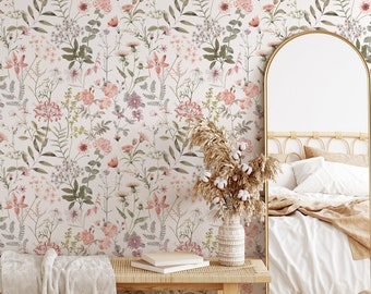May include: A bedroom interior with a floral wallpaper featuring a variety of pink and green flowers. A wooden bench with a white throw blanket sits in front of the wallpaper. A wicker basket and a vase with dried flowers are on the floor. A large mirror with a gold frame is on the wall. A bed with a white and brown blanket is in the background.