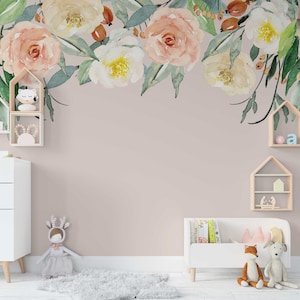 May include: A light pink wall with a floral garland made of watercolor flowers in shades of pink, white, and yellow. The garland is hanging above a white dresser and a white toy bench with books and stuffed animals.