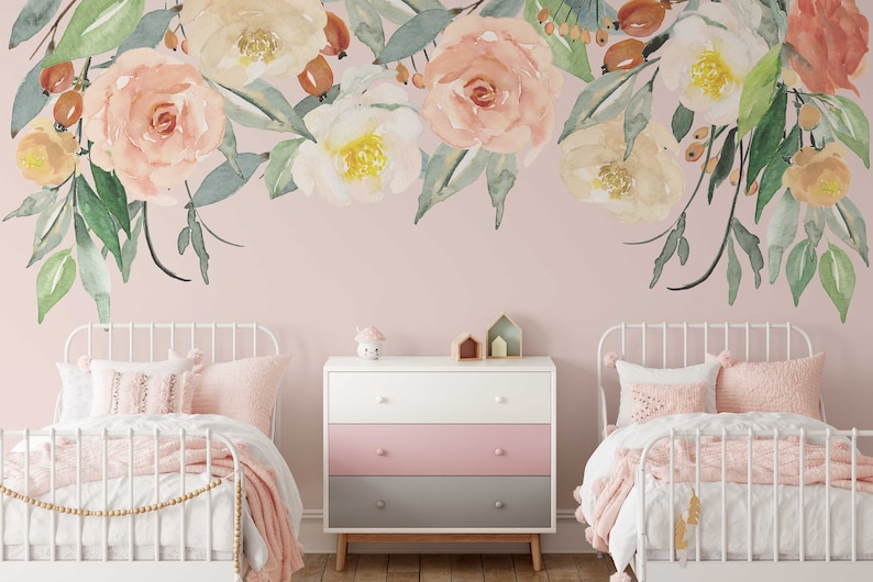 May include: Two white twin beds with pink bedding and a white dresser with three drawers. The dresser has a pink, white, and gray color scheme. The wall behind the beds is a light pink color and features a floral decal with watercolor-style flowers in shades of pink, white, and green.