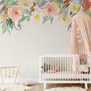 May include: A white crib with a pink and green patterned blanket and a pink canopy with white clouds hanging from it. The crib is in a nursery with a floral wallpaper and a wooden toy stand.