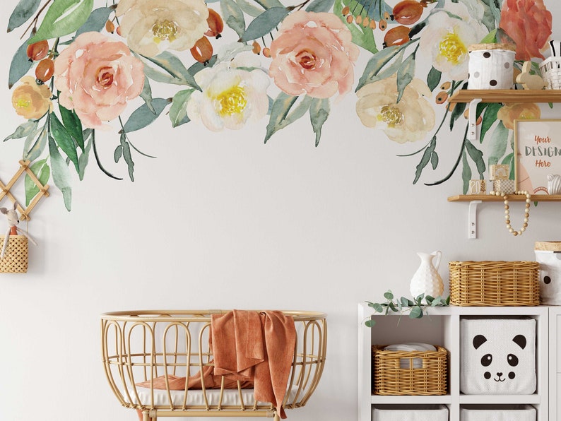 May include: A white wall with a floral decal featuring watercolor pink and white flowers with green leaves. A wicker bassinet with a burnt orange blanket is in the foreground. A white shelf with wicker baskets and a panda-themed storage bin is on the right side of the image.