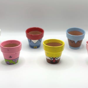 May include: Six small, hand-painted terracotta flower pots. Each pot is decorated with a different cartoon character design, featuring bright colors like pink, yellow, blue, and red. The pots are empty and ready for planting.