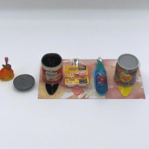 May include: Miniature food items including a can of Boyardee, a Lunchables box, a blue soda bottle, and a Chupa Chups can. A small orange food item and a gray can lid are also present. The items are arranged on a decorative surface.