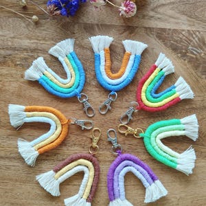 Puede incluir: Una colección de llaveros de arcoíris de macramé hechos a mano en varios colores, incluyendo azul, naranja, rojo y morado. Cada llavero presenta un cierre metálico y un detalle de flecos. Los llaveros están dispuestos sobre una superficie de madera, con flores secas en el fondo.