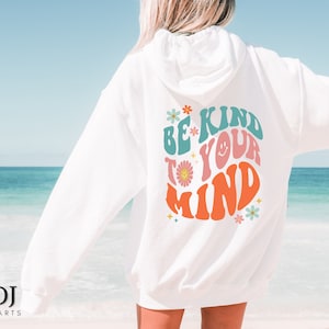 Puede incluir: Sudadera con capucha blanca con el texto "BE KIND TO YOUR MIND" en un estilo retro. El diseño incluye una cara sonriente y detalles florales en verde azulado, rosa y naranja. Prenda de vestir informal.