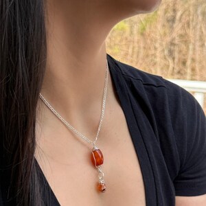 May include: A silver chain necklace with a large, orange-red gemstone pendant. The pendant is wire-wrapped and has a smaller, matching gemstone dangling below. The necklace is worn against a black top.