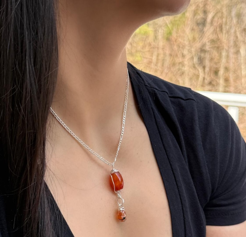 May include: A silver chain necklace with a large, orange-red gemstone pendant. The pendant is wire-wrapped and has a smaller, matching gemstone dangling below. The necklace is worn against a black top.
