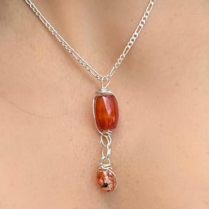 May include: A silver necklace featuring a pendant with two gemstone beads. The top bead is a large, orange-red, barrel-shaped stone. The bottom bead is a smaller, speckled, oval-shaped stone. The beads are connected by silver wire.