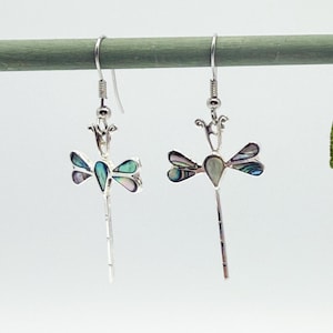 Könnte beinhalten: Silberne Libellenohrringe mit schillernden Abalone-Flügeln und langen, schlanken Schwänzen. Die Ohrringe haben ein detailliertes Design mit einem blütenartigen Element an der Oberseite. Die Libellenflügel zeigen Farbtöne von Blau, Grün und Lila.