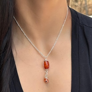 May include: A silver chain necklace with two gemstone pendants. The top pendant is a reddish-orange, barrel-shaped stone, and the bottom pendant is a smaller, speckled stone. The stones are wire-wrapped.