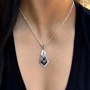 May include: A silver pendant necklace with a geometric design. The pendant has layered, diamond-shaped elements with black and iridescent accents. The necklace is worn on a silver chain against a dark background.