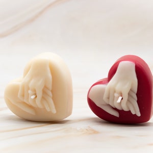 May include: Two heart-shaped candles with a couple's hands holding each other. One candle is white and the other is red.
