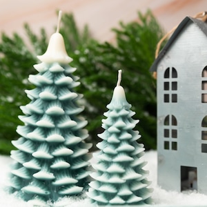 Puede incluir: Dos velas de árbol de Navidad azul y blanco y una casa de metal con ventanas recortadas. Las velas y la casa están sobre una superficie blanca que parece nieve.
