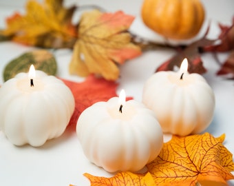 May include: Three white pumpkin-shaped candles with lit wicks, surrounded by colorful autumn leaves and small pumpkins. The candles are the focal point, evoking a seasonal, fall theme. The leaves are in shades of orange, yellow, and red.