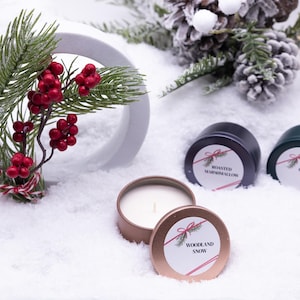 May include: A festive holiday scene featuring three scented candles in tins, with labels reading "Woodland Snow," "Roasted Marshmallow," and "Fraser Fir." The candles are surrounded by artificial snow, pine branches, and red berries.