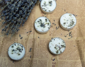 Op de afbeelding: Vijf witte theelichtjes met gedroogde lavendelbloemen die in de was zijn ingebed. De kaarsen zijn gerangschikt op een bruin papieroppervlak met een takje lavendel op de achtergrond.
