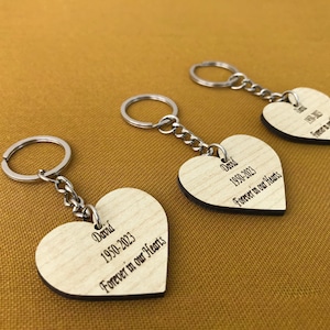 Könnte beinhalten: Drei silberne Schlüsselanhänger mit herzförmigen Anhängern aus Holz. Jeder Anhänger trägt eine personalisierte Inschrift mit einem Namen, Daten und dem Satz "Forever in our Hearts".