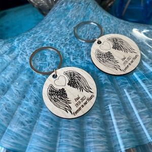 May include: Two round wooden keychains with silver keyrings. Each keychain features a heart and wing design, with the text "Paul 1958-2025 Forever in our Hearts" engraved in black. The keychains are on a blue surface.