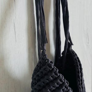 May include: A dark charcoal gray handbag crafted from thick, chunky fabric yarn. The bag features a textured, crocheted or knitted stitch pattern and includes two long, flat fabric straps that are knotted at the top for hanging purposes.