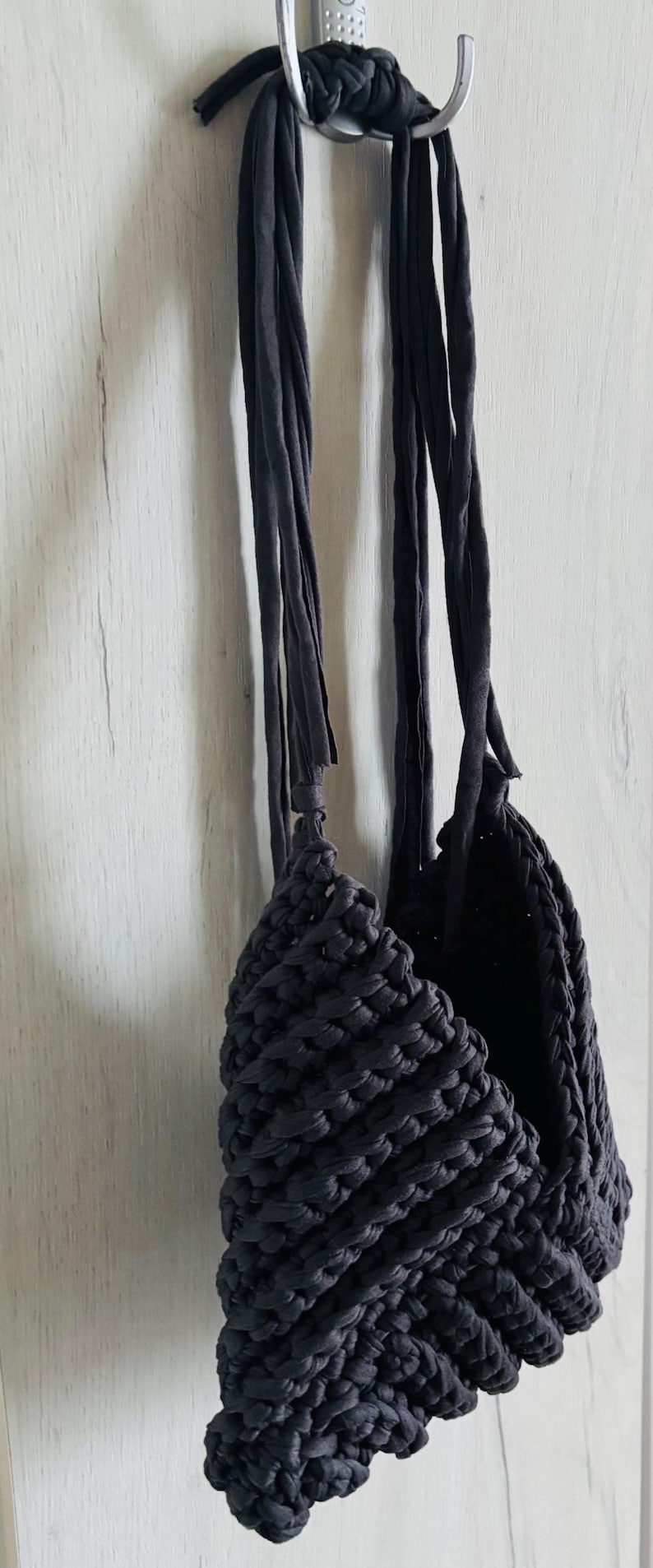 May include: A dark charcoal gray handbag crafted from thick, chunky fabric yarn. The bag features a textured, crocheted or knitted stitch pattern and includes two long, flat fabric straps that are knotted at the top for hanging purposes.