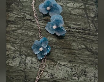 May include: A delicate necklace and matching earrings set featuring blue hydrangea petals accented with small white pearls. The jewelry is crafted with a thin metallic chain. The text @dechicreazioni is visible at the bottom of the image.