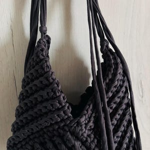 Pode incluir: Uma bolsa de mão cinza-carvão escuro feita de tiras de tecido grossas e trançadas. A bolsa apresenta um padrão de tricô diagonal texturizado com uma abertura profunda em forma de V e longas tiras de tecido pendentes nas laterais.