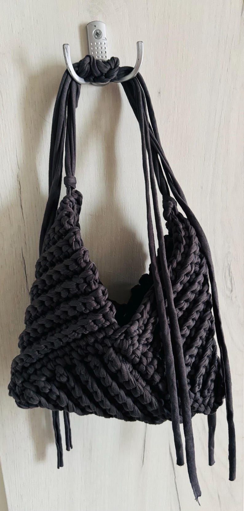 May include: A dark charcoal gray handbag crafted from thick, braided fabric strips. The bag features a textured, diagonal knit pattern with a deep V-shaped opening and long, dangling fabric straps hanging from the sides and top handle.