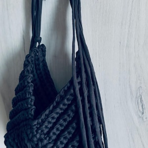 May include: A dark charcoal gray crochet bag crafted from thick fabric yarn. The bag features a textured, ribbed stitch pattern and long, thin fabric straps that extend from the top opening, hanging down the side of the bag structure.