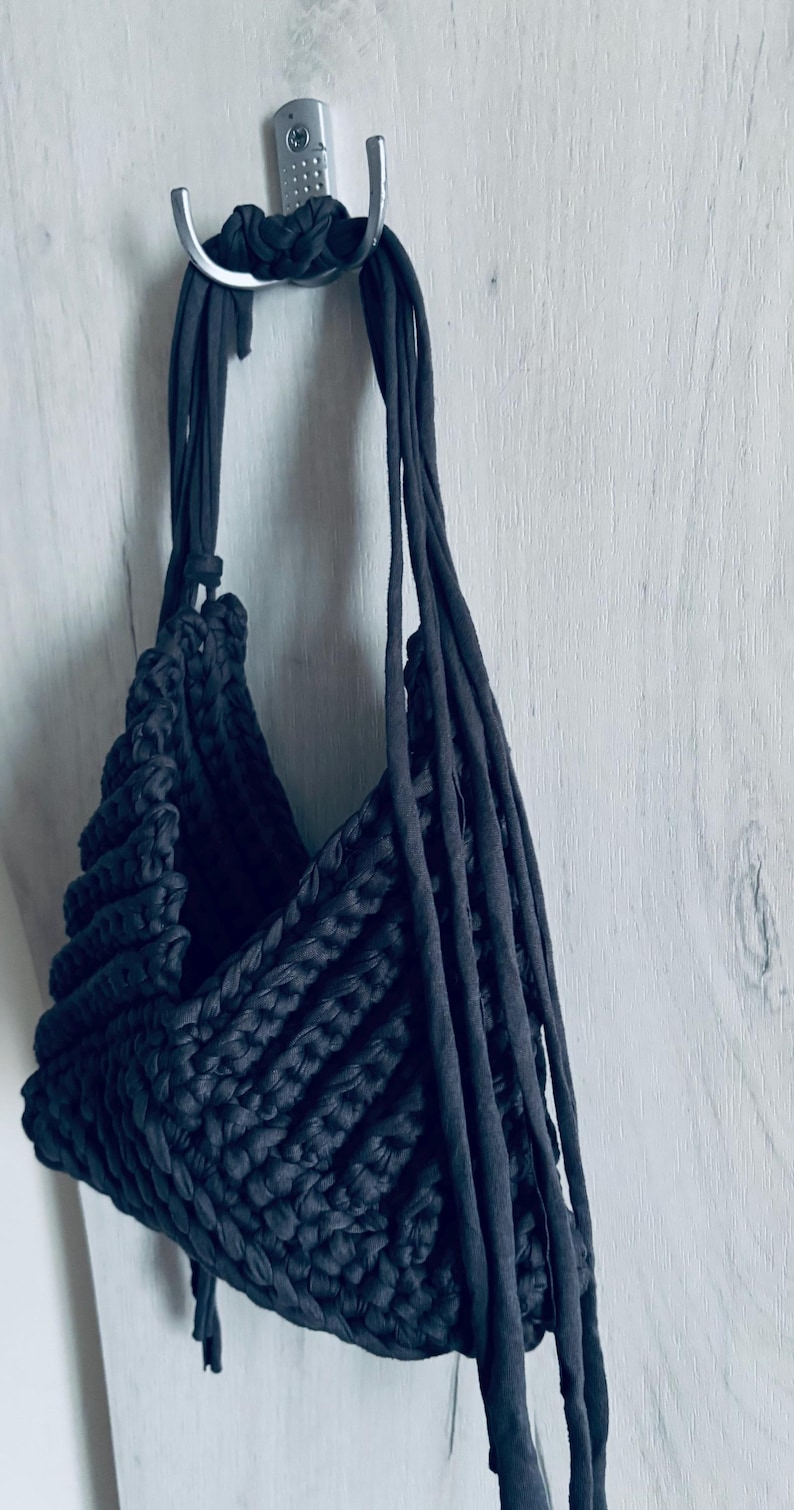 May include: A dark charcoal gray crochet bag crafted from thick fabric yarn. The bag features a textured, ribbed stitch pattern and long, thin fabric straps that extend from the top opening, hanging down the side of the bag structure.