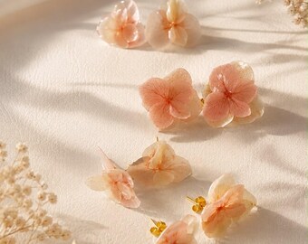 May include: A collection of stud earrings featuring delicate, preserved hydrangea petals in soft shades of peach and cream. Each earring is attached to a small gold-toned metal post, showcasing the natural, translucent texture of the flower petals.