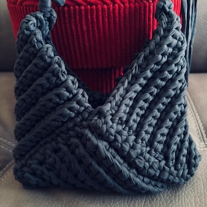 May include: A dark gray crochet handbag crafted from thick fabric yarn, featuring a textured chevron pattern and two knotted top handles. The bag has a V-shaped opening and a structured, woven appearance with visible loops throughout the design.