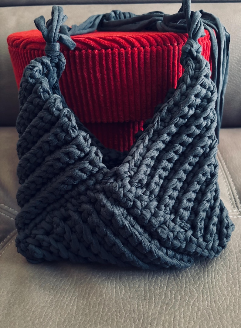 May include: A dark gray crochet handbag crafted from thick fabric yarn, featuring a textured chevron pattern and two knotted top handles. The bag has a V-shaped opening and a structured, woven appearance with visible loops throughout the design.