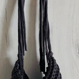 May include: A dark charcoal gray handbag crafted from thick, braided fabric cord. The bag features a triangular, textured knit pattern and two long, slender fabric straps that extend upward, allowing the item to be suspended from a metal hook.