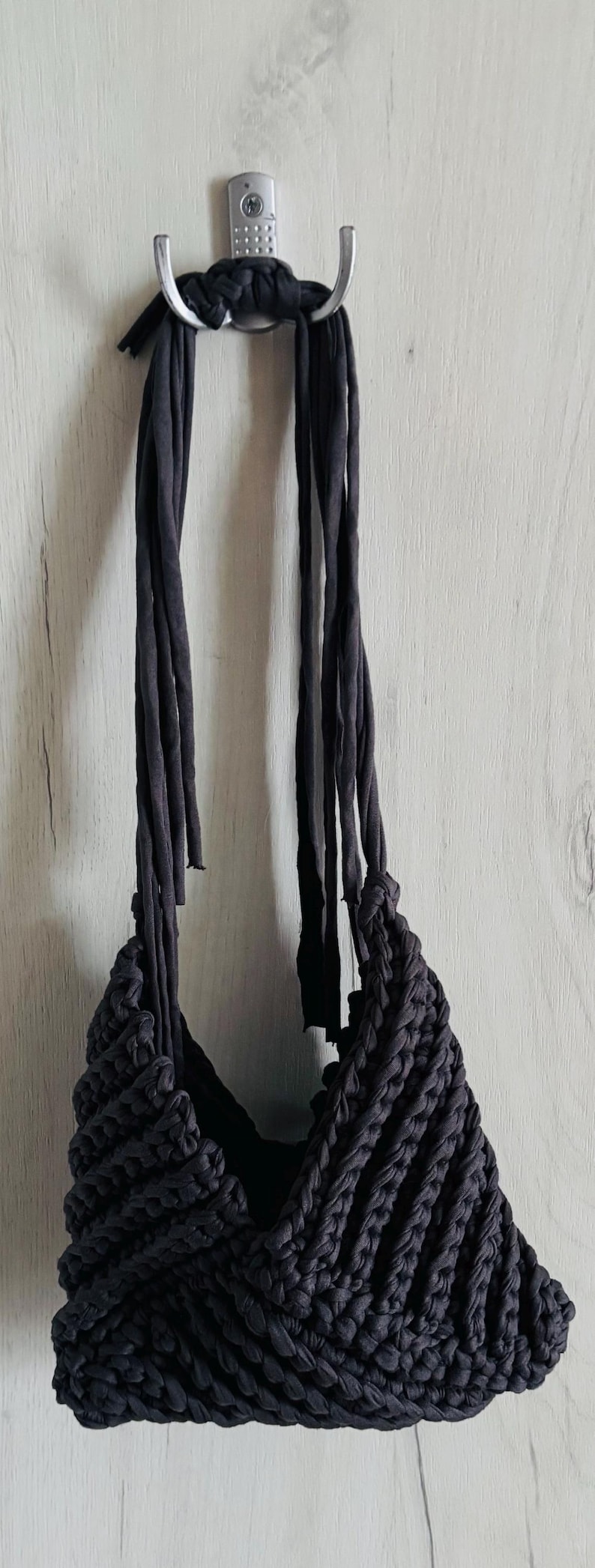 May include: A dark charcoal gray handbag crafted from thick, braided fabric cord. The bag features a triangular, textured knit pattern and two long, slender fabric straps that extend upward, allowing the item to be suspended from a metal hook.