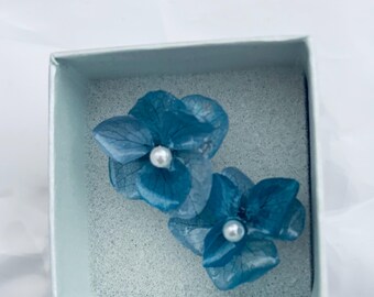 May include: A pair of blue hydrangea flower earrings, each featuring delicate, translucent petals and a small, round white pearl at the center. The earrings are presented inside a square, light-colored box with a textured foam insert.