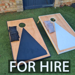 May include: Two wooden cornhole boards with a natural wood finish and painted geometric designs in black and light blue. Black and gray bean bags are placed on the boards. The text "FOR HIRE" is displayed at the bottom.