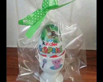 May include: A white ceramic egg cup with a colorful design holds a Kinder Surprise chocolate egg wrapped in clear plastic. A green ribbon with white polka dots is tied around the top of the plastic wrap.