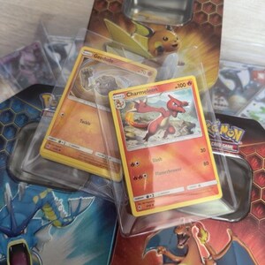 May include: Three Pokemon Trading Card Game tins with various Pokemon cards inside. The tins are orange, blue and yellow. The cards feature Charmeleon, Geodude and a blue Pokemon.