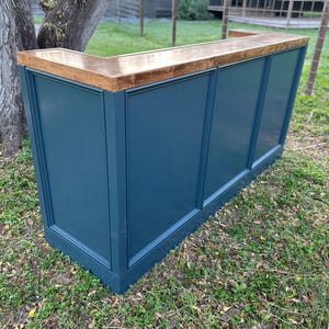 May include: A blue wooden bar with a light brown countertop. The bar has a rectangular shape with paneled sides. It is outdoors on a grassy area, near a tree.