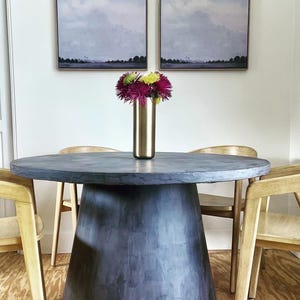 May include: A round, dark gray table with a pedestal base, surrounded by four wooden chairs. A gold vase holds a bouquet of purple, green, and yellow flowers. Two framed landscape paintings hang on the white wall in the background.