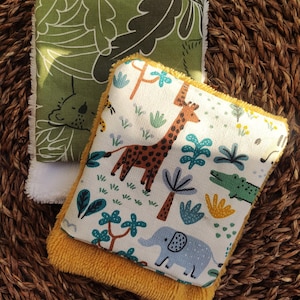 Puede incluir: Dos cuadrados reutilizables. Uno es verde con un estampado de hojas blancas, el otro tiene un borde amarillo y un fondo blanco con un estampado de jirafa, elefante y cocodrilo. Los cuadrados están sobre una superficie tejida marrón.