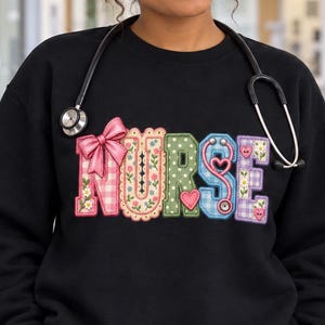 May include: A black long-sleeve sweatshirt featuring the word NURSE in large, multi-coloured, patterned block letters. The letters include floral, gingham, and polka dot designs with a pink bow and heart accents. A stethoscope rests around the neck.