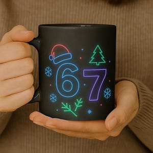 Op de afbeelding: Zwarte keramische mok met een neon ontwerp met de cijfers 6 en 7, een kerstmuts, sneeuwvlokken en een kerstboom. De mok heeft een handvat en wordt door een persoon vastgehouden.