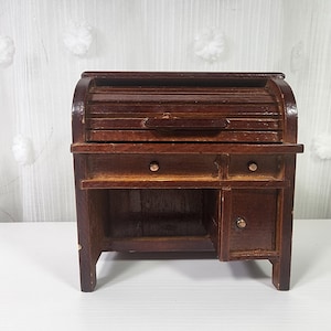 May include: A miniature dark brown wooden roll-top desk featuring a curved sliding top, one central drawer, one smaller side drawer, and a small cabinet door on the right side. Each drawer and the cabinet door are fitted with a small, round metal knob.