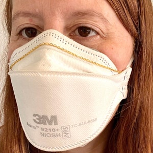 May include: A white 3M Aura 9210+ N95 respirator mask with a gold chain attached to the ear loops. The mask has the text "3M Aura 9210+ N95 TC-84A-6669" printed on it.