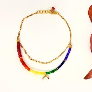 May include: A gold-colored necklace with two strands. The lower strand features a rainbow of colorful beads, with a small rainbow charm. The upper strand is a simple gold chain. A dried leaf is next to the necklace.