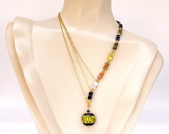 May include: A gold-toned necklace with a pendant and colorful beads. The pendant is a yellow circle with black detailing. The necklace features a layered design with a variety of square and rectangular beads in shades of pink, orange, and black.