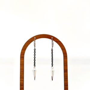 May include: A pair of silver-toned earrings with a black chain and a cone-shaped pendant. The earrings are displayed on a wooden arch-shaped stand against a white background. The earrings have a modern, edgy design.