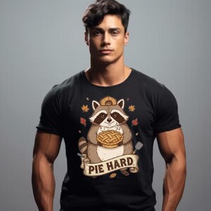 Könnte beinhalten: Schwarzes T-Shirt mit einem Cartoon-Waschbären, der einen Kuchen hält. Der Waschbär trägt einen Hut und ist von Herbstblättern umgeben. Der Text "PIE HARD" ist auf einem Banner darunter zu sehen.
