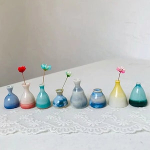 May include: A collection of eight small, colorful ceramic vases, each holding a single artificial flower. The vases are in shades of blue, pink, yellow, and green. The arrangement is set on a white lace surface.